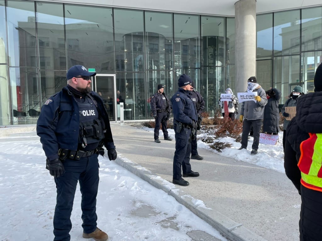 Police standing outside the Qaumajuq section of the Winnipeg Art Gallery in winter, pro-Palestinian demonstrators hold “re-fund UNRWA” and other signs